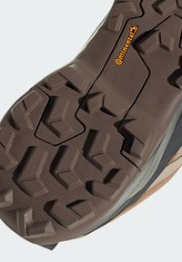 Outsole butów zewnętrznych cechuje agresywny wzór bieżnika z wielokierunkowymi kołkami w ciemnobrązowej gumie. Teksturowana powierzchnia dla lepszej przyczepności.