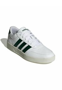 Zapatillas deportivas blancas con rayas verdes y logo, hechas de cuero sintético, con puntera redondeada y suela texturizada. Diseño estándar con cordones.