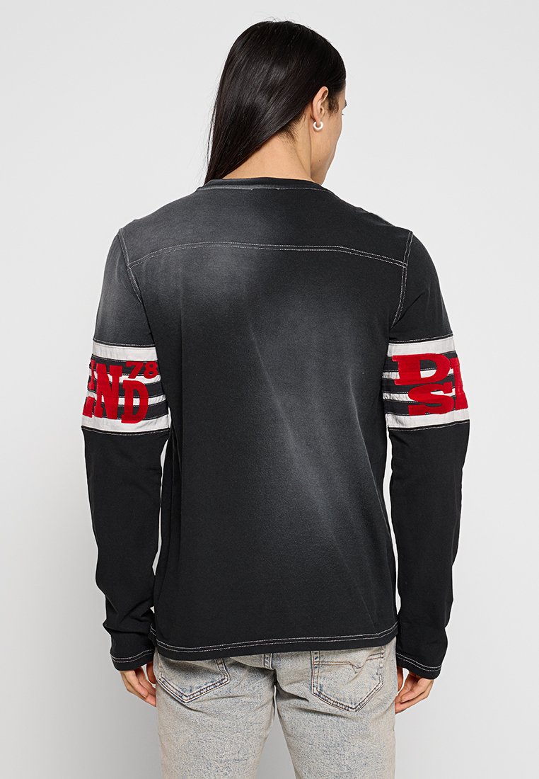 Schwarzes Langarmshirt mit roten und weißen Streifenakzenten an den Ärmeln, mit auffälligem Schriftzug am linken Arm. Mit abgenutzter Textur.