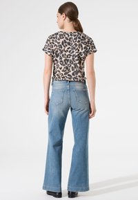 Camiseta de estampado de leopardo en beige y negro, combinada con jeans acampanados de color azul claro. El conjunto presenta un corte relajado y bolsillos traseros visibles.