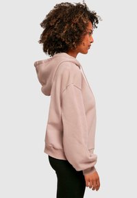 Lichtroze hoodie met een relaxte pasvorm, oversized mouwen, geribbelde manchetten en een trekkoordkap, weergegeven tegen een neutrale achtergrond.