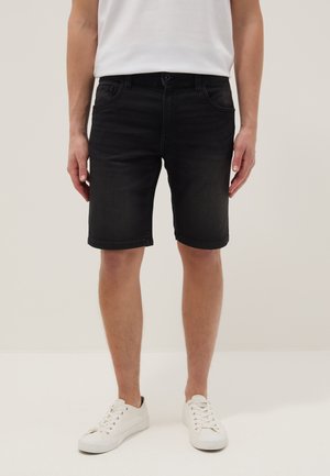 Man die zwarte denim shorts, een wit T-shirt en witte sneakers draagt, staand tegen een effen lichte achtergrond.