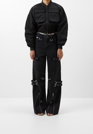 Person wearing black cropped jacket with front pockets and high-waisted black pants featuring multiple rings, straps, and front zippers.