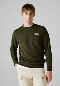 Olijfgroen sweatshirt met lange mouwen, ronde hals en het "RED BULL RACING" logo in het wit. Licht textuur en geribbelde manchetten en onderkant.