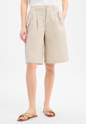 Beige knielange op maat gemaakte shorts met knoopsluiting op de zijzakken, gedragen met beige gesnoerde slides.