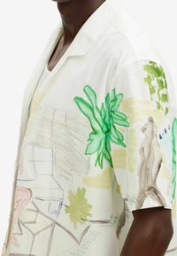 Camicetta a maniche corte realizzata in tessuto bianco con colorate decorazioni tropicali dipinte a mano, con accenti verdi, beige e blu delicato.