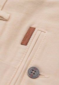 Tissu beige présentant une texture à côtes horizontales, avec une étiquette en cuir et un accent de bouton en métal. Comprend un détail de poche latérale.