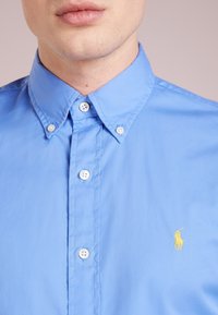 Světle modrá košile na knoflíky vyrobená z bavlny. Má klasický límec, bílé knoflíky a malé žluté polo logo na levé části hrudníku.