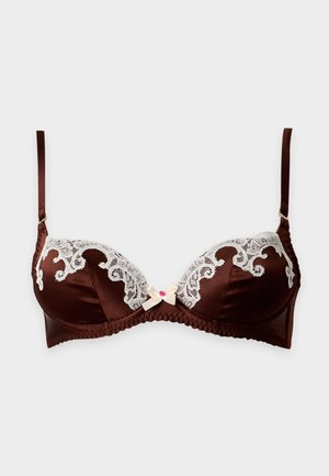Femme aux longs cheveux foncés portant une lingerie marron avec des détails en dentelle blanche, debout pieds nus sur un fond blanc uni.