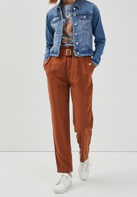 Veste en denim bleue, avec des boutons en argent et deux poches poitrine. Pantalons marron ajustés avec une jambe large et une ceinture, associés à des baskets blanches.