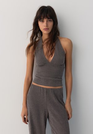 Femme aux longs cheveux bruns portant un haut gris texturé dos-nu et un pantalon large assorti, se tenant devant un fond clair uni.