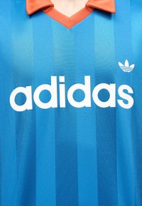 Μπλε αθλητική μπλούζα Adidas με κάθετες ρίγες, λευκό κείμενο λογότυπου Adidas, έμβλημα τριφύλλι και πορτοκαλί γιακά.