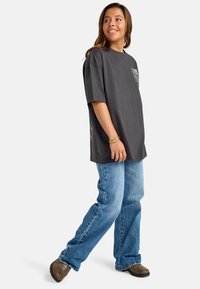 T-shirt grafico in cotone grigio scuro, con tasca decorata da un design colorato. Abbinato a jeans a gamba dritta blu chiaro e scarpe marroni.