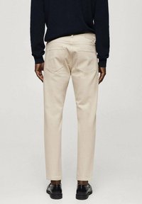 Pantalones beige de algodón, con corte recto, dos bolsillos traseros con solapas en ángulo y tobillos rematados. Combinados con zapatos negros.