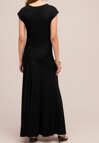 Femme portant une robe noire longue avec des manches courtes et des côtés froncés, accessoirisée de bracelets en or et de sandales à talons noires.