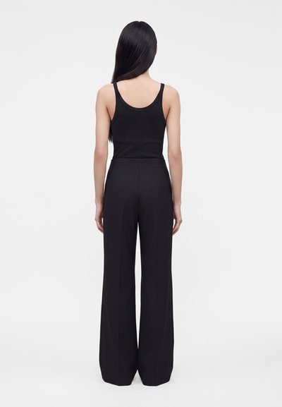 Femme aux longs cheveux noirs porte un haut noir sans manches et un pantalon noir taille haute à jambes larges, debout de dos sur un fond blanc.