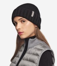 Svart ribbad mössa med uppvikt kant och en liten sydd etikett. Bärs med en grå pufferväst och en figurnära svart polotröja. Slät textur.