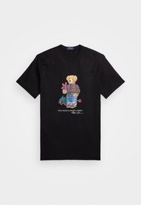 Černé tričko s medvídkem v úpletu s vzorem a džínách, který drží zabalený dárek, s nápisem "Polo Bear by Ralph Lauren" dole.
