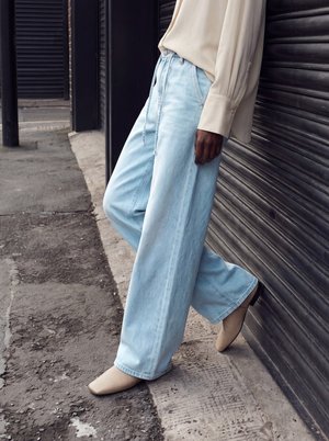 Personne portant un jean large bleu clair, un chemisier beige et des chaussures beiges à bout carré, debout devant un volet métallique noir à l'extérieur.