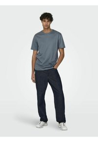 T-shirt grigio a maniche corte realizzato in tessuto morbido, abbinato a pantaloni in denim blu scuro. Il modello indossa scarpe da ginnastica bianche, in piedi su uno sfondo neutro.