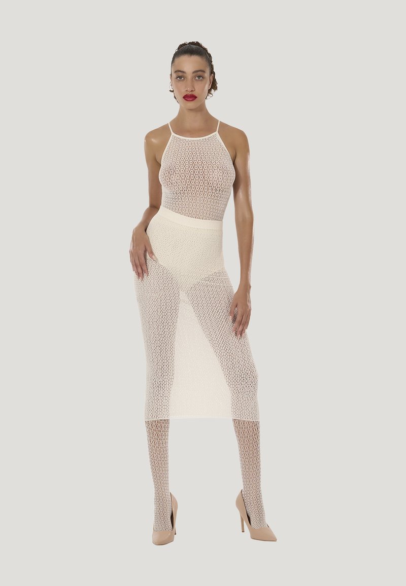 Vrouw met donker haar in een knot, gekleed in een doorschijnende witte mesh jurk en beige hoge hakken, staand met één hand op haar heup tegen een effen achtergrond.