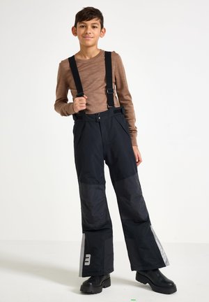 Pantaloni da neve neri con bretelle nere regolabili, vita elastica e dettagli riflettenti sui lati. Indossati con una maglietta a maniche lunghe marrone chiaro.