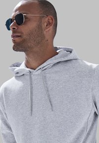 Haut à capuche gris clair avec un cordon de serrage et une texture douce. Porté avec des lunettes de soleil sombres, présentant un design élégant et des accents en métal.
