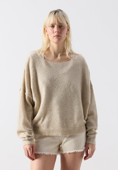 Femme blonde portant un pull beige ample et un short en jean clair effiloché, debout devant un fond blanc uni.