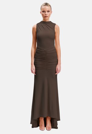 Frau steht aufrecht und trägt ein ärmelloses dunkelbraunes High-Low-Kleid mit gerafften Details an der Taille und transparente offene Zehensandalen.