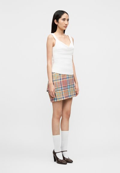 Λευκή αμάνικη μπλούζα με V λαιμόκοψη, σε συνδυασμό με πολύχρωμη καρό μίνι φούστα. Το μοντέλο φοράει λευκές κάλτσες μέχρι το γόνατο και καφέ παπούτσια με τακούνι.