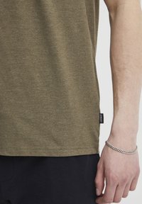 Olivengrünes T-Shirt mit kurzen Ärmeln aus weichem, strukturiertem Stoff. Der Saum hat ein schwarzes Etikett. Eine Hand ruht auf einer dunklen, gerippten Oberfläche.