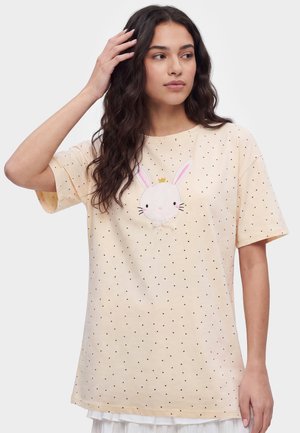 Femme aux longs cheveux foncés portant un t-shirt jaune clair avec de petits cœurs noirs et un motif de lapin blanc avec une couronne sur la poitrine.
