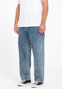 Vaqueros baggy de denim deslavado en azul claro con una ligera textura desgastada. Presentan un bolsillo lateral y un diseño de pierna recta, combinados con zapatillas negras.