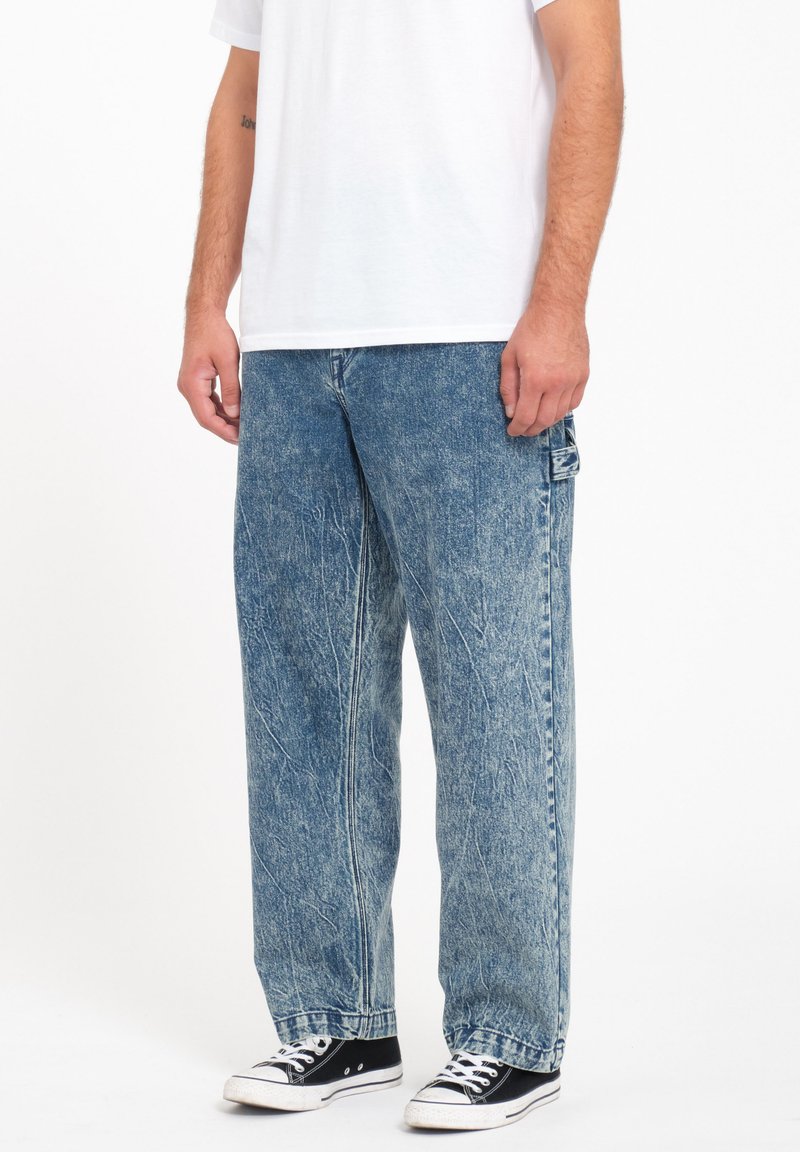 Vaqueros baggy de denim deslavado en azul claro con una ligera textura desgastada. Presentan un bolsillo lateral y un diseño de pierna recta, combinados con zapatillas negras.
