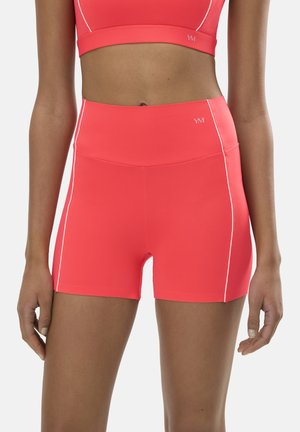 Mujer con pantalones deportivos ajustados de talle alto en color coral con ribetes blancos y sujetador deportivo a juego, de pie con los brazos relajados a los lados.