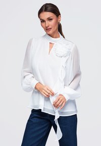 Femme portant un chemisier blanc transparent avec un détail floral et un lien volanté, associé à un jean bleu foncé, posant devant un fond clair uni.