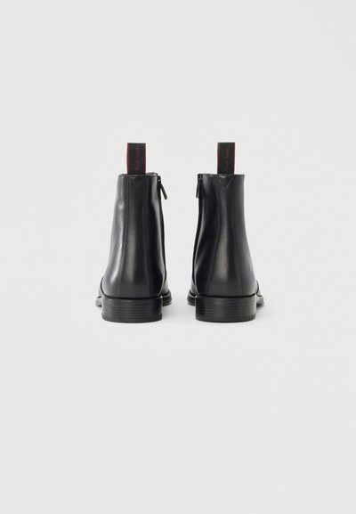 Botines de cuero negro con acabado liso, cierre de cremallera en la parte trasera y detalle de lengüeta de tirador rojo en contraste en el talón.