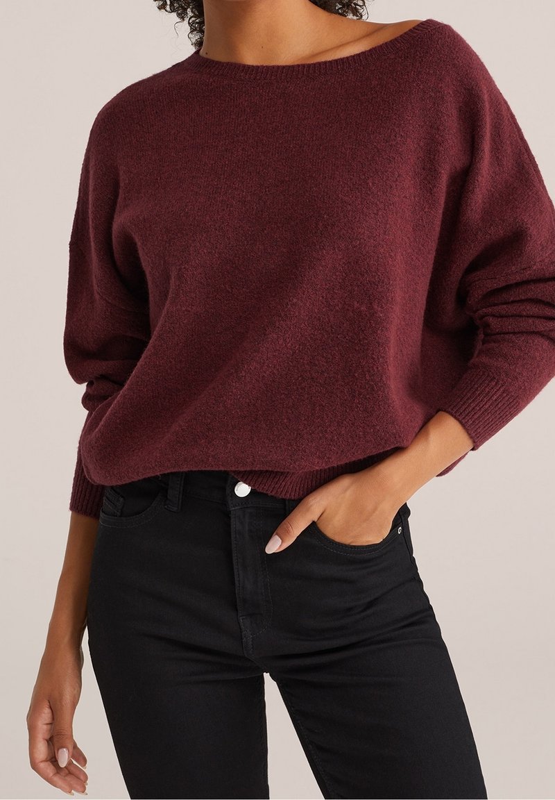 Vínový pletený svetr se širokým výstřihem a žebrovanými manžetami, doplněný o přiléhavé černé džíny, které zdůrazňují měkkou texturu a ležérní střih.