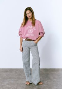 Mujer luciendo un sudadera rosa con "Authentic Brownie", pantalones de pierna ancha a rayas, cinturón marrón y sandalias, de pie contra una pared blanca.