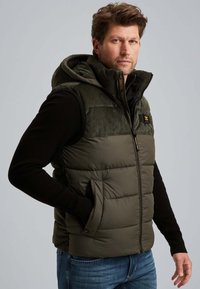 Homme portant une veste sans manches rembourrée verte avec capuche, sur une chemise à manches longues noire et un jean bleu, se tenant avec une main dans la poche.
