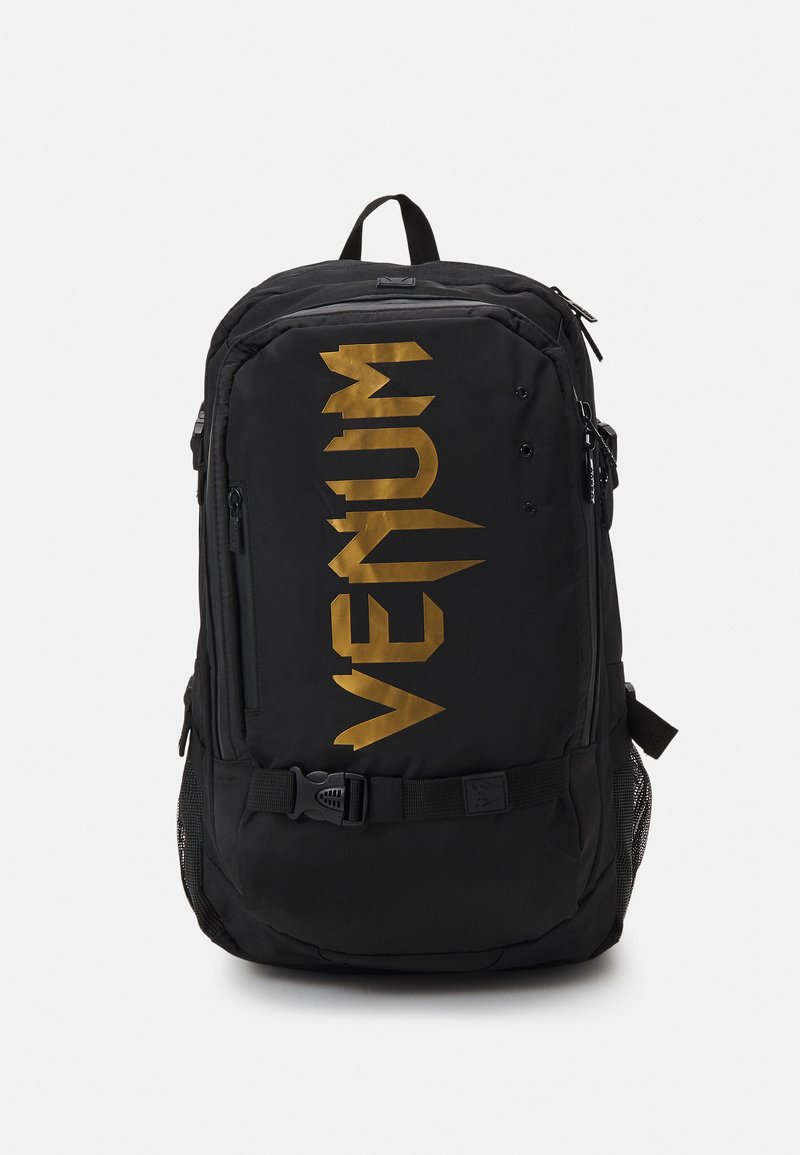 Črn nahrbtnik z izrazitim zlatim logotipom "VENUM", stranskimi mrežastimi žepi, prilagodljivimi naramnicami in trdim zapiranjem s sponko.