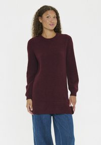 Vrouw in een bordeauxrode sweater met lange mouwen en losse blauwe spijkerbroek, staand tegen een effen witte achtergrond.