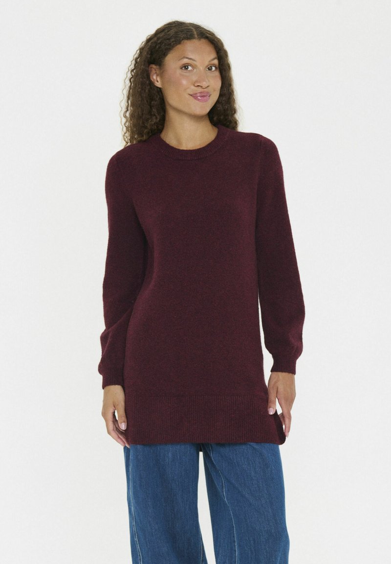 Vrouw in een bordeauxrode sweater met lange mouwen en losse blauwe spijkerbroek, staand tegen een effen witte achtergrond.