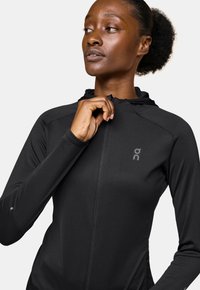 Schwarze Langarm-Zip-Hoodie mit strukturiertem Finish, dehnbarem Stoff und einer enganliegenden Kapuze; mit einem Logo auf der Brust und enganliegenden Handgelenken.