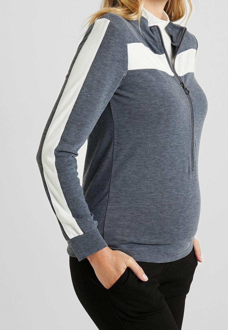 Sweat-shirt zippé gris avec des rayures blanches sur les épaules, manches longues ajustées et tissu texturé. Poches latérales visibles au niveau des hanches.