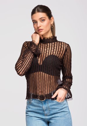 Nainen, jolla on vedetyt hiukset, pukeutuneena läpikuultavaan mustaan pitsipaitaan mustan rintaliivin yllä ja sinisiin farkkuihin, poseeraa yhdellä kädellä lähellä kasvoja ja toinen käsi taskussa.