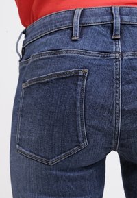 Vaqueros de mezclilla azul oscuro con un diseño clásico de cinco bolsillos. Presentan costuras visibles y un leve desgaste en la tela. Detalle del bolsillo trasero mostrado.