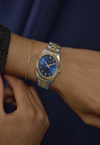 Încheietura cu un ceas Timex din metal bicolor, cu cadran albastru și accente aurii, alături de o brățară delicată din aur, pe o piele de culoare închisă.