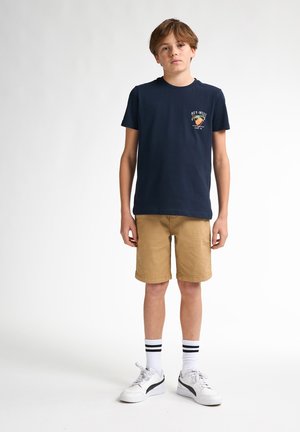 T-shirt di cotone navy con logo, pantaloni corti beige, sneakers bianche con calze a strisce nere. Stile semplice, vestibilità rilassata e casual.