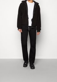 Chaqueta de forro polar negra con capucha, cierre frontal y puños acanalados. Llevada sobre una camiseta blanca y pantalones negros. Zapatillas negras con superficie texturizada.